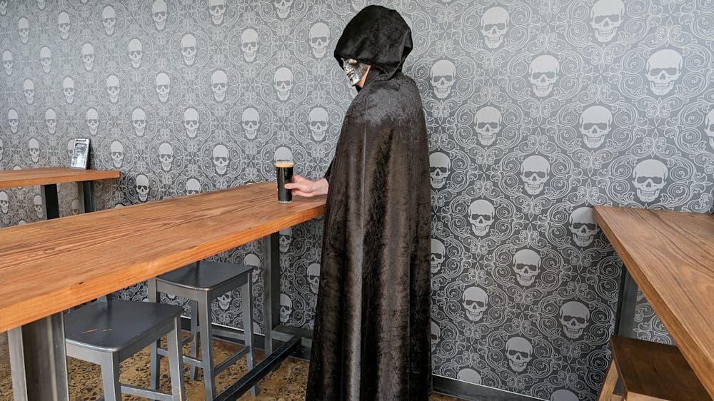Grim reaper character standing with a glass of dark beer in hand. Wallpaper behind is of skulls