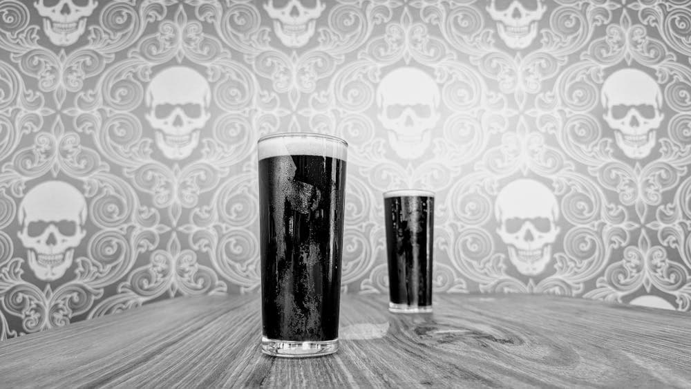 Two glasses of dark beer staggered with skull wallpaper out of focus behind. Image is in greyscale
