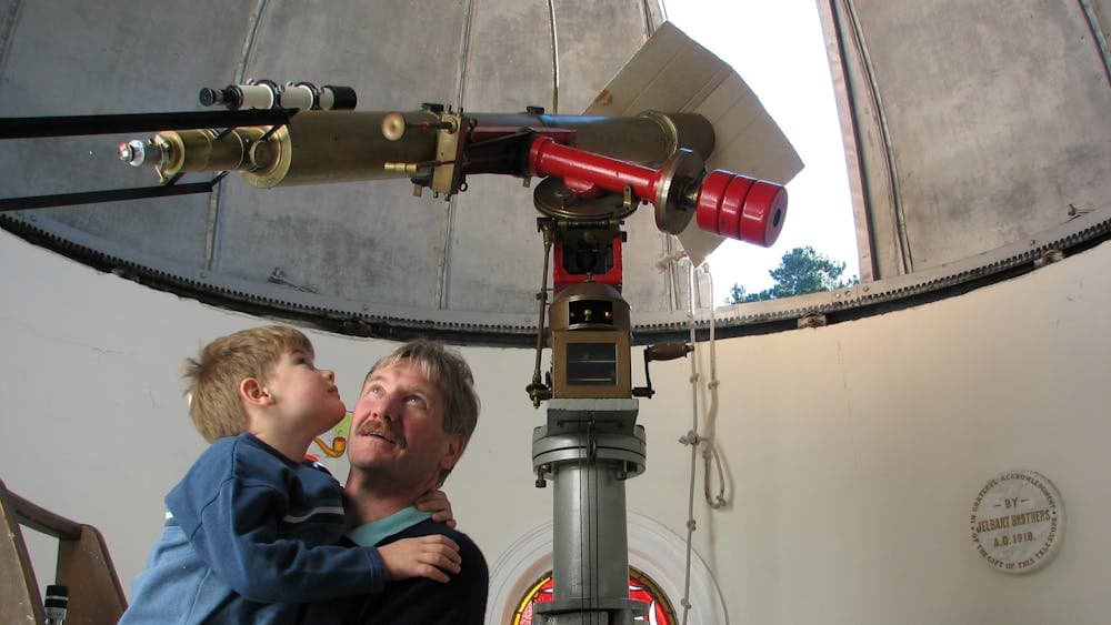 adult holding child up to look at 5 inch brass refractor telescope in dome with roof open