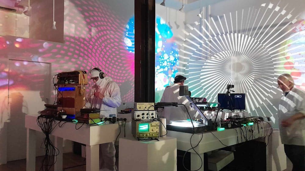 Men in white coats playing analogue synthesizers with lights lit up in psychadelic colours.