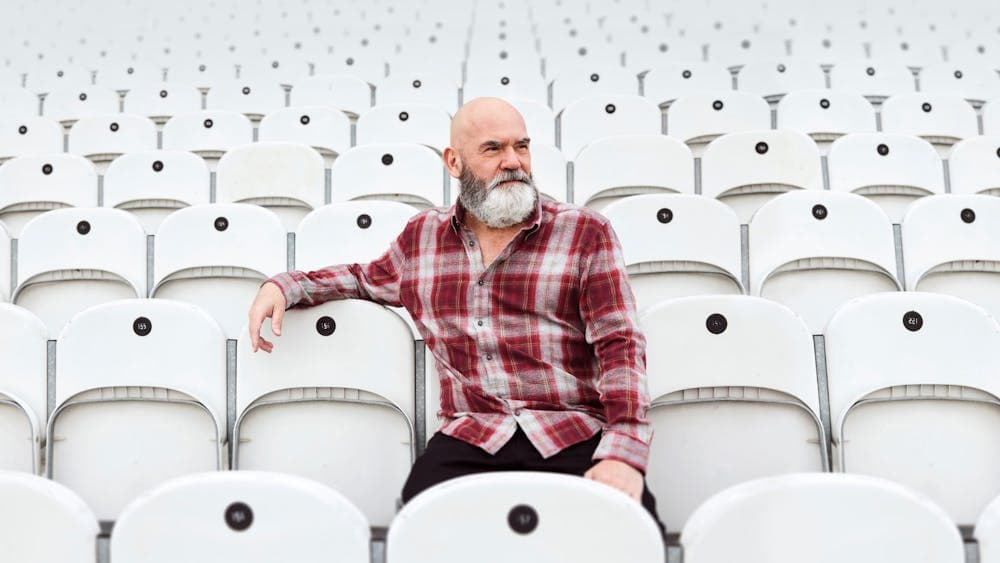 Marty Sheargold in a flannel shirt seated in a stadium with white chairs