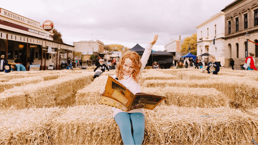 Girl sitting on hay bale reading a book with one arm in the air