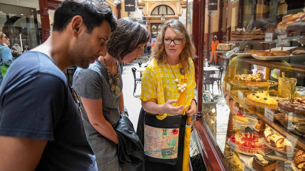 Guide wearing a branded tour bag and yellow top, points oto cakes in a window to 2 tour guests