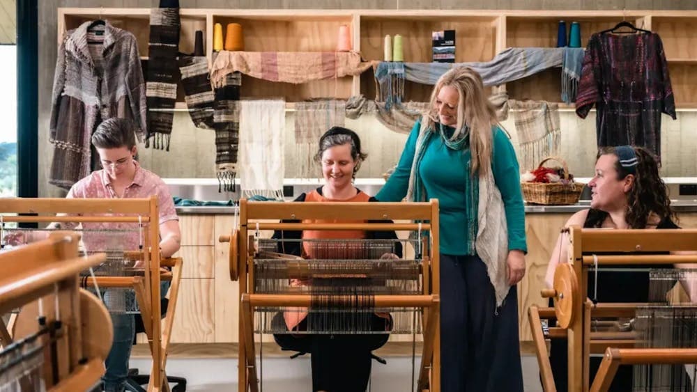 Three women seated at looms with instructor standing between them inside workshop