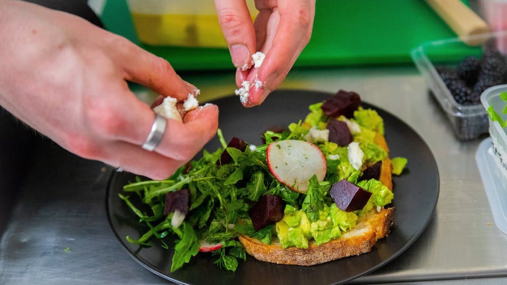 Smashed Avocado being plated in the kitchen with hands crumbling goat's cheese over the top