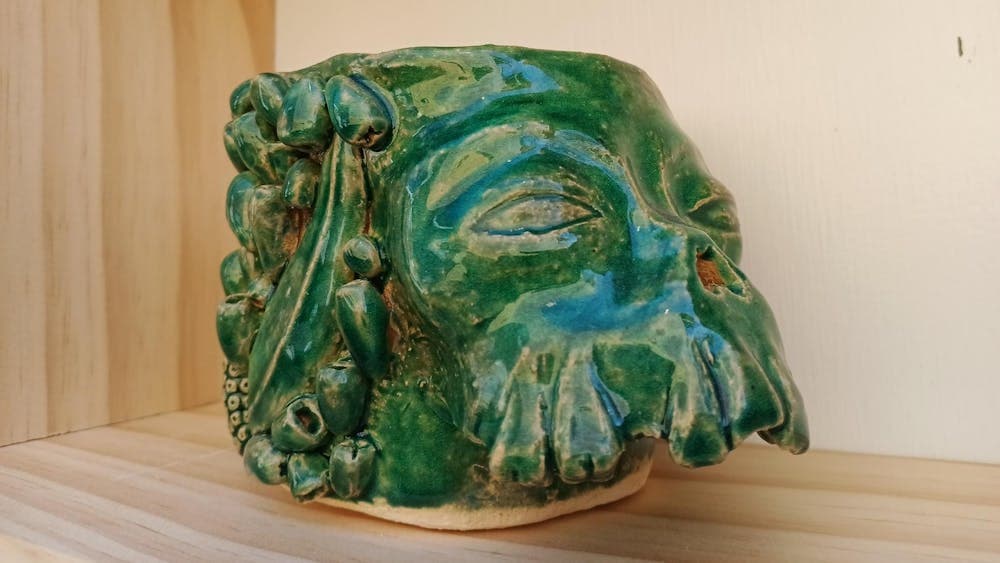 Ceramic sculpture of a morphing skull painted in green glaze on a wooden shelf.