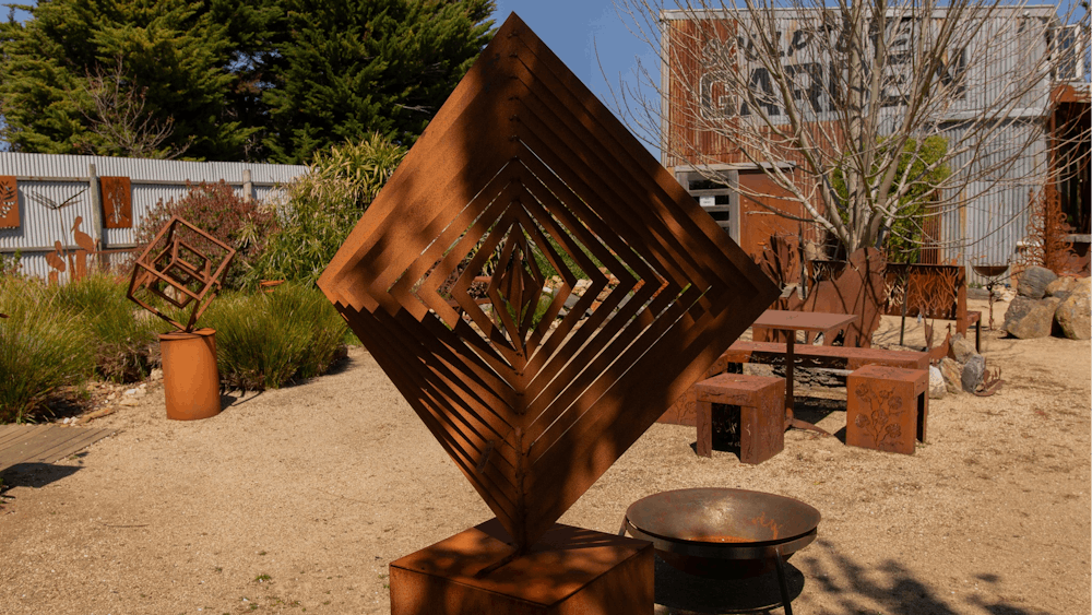 A metal square sculpture in the garden with bench seats and fire pit behind.