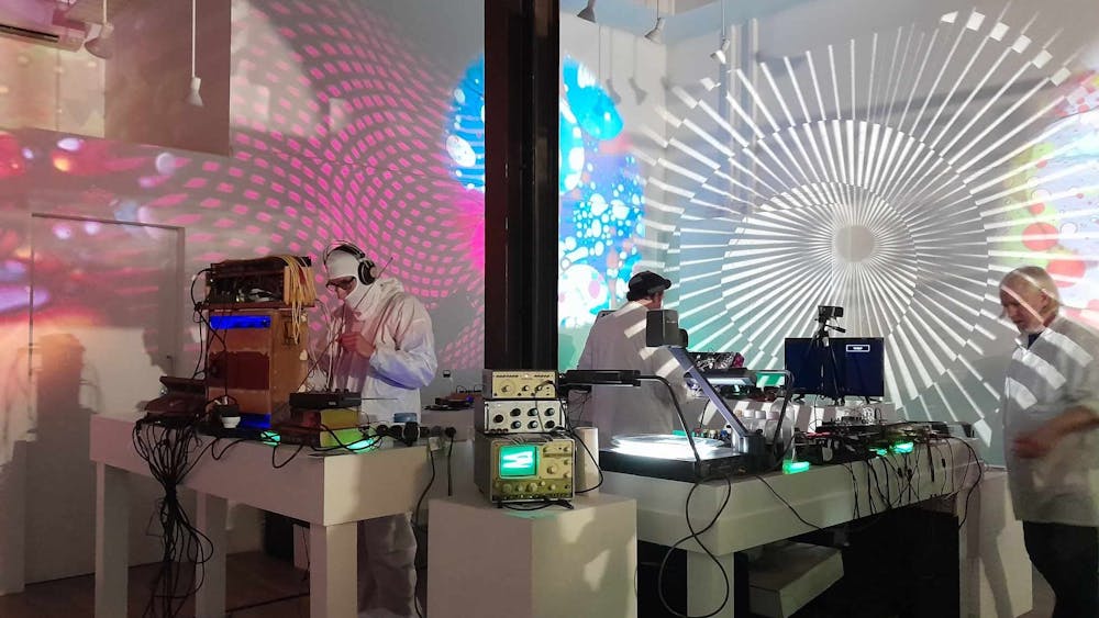 Men in white coats playing analogue synthesizers with lights lit up in psychadelic colours.