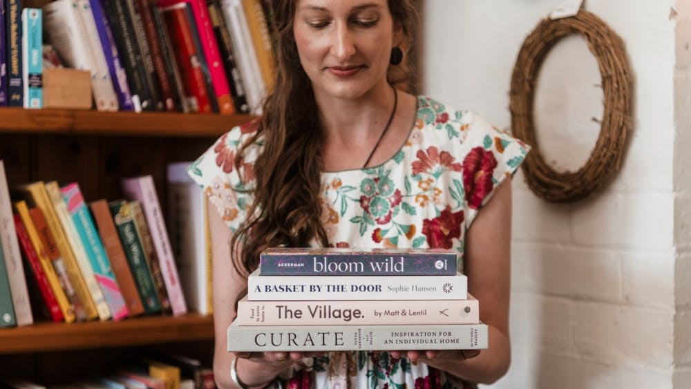 a woman looking down in a floral dress holding a stack of books, standing in front of bookcase.