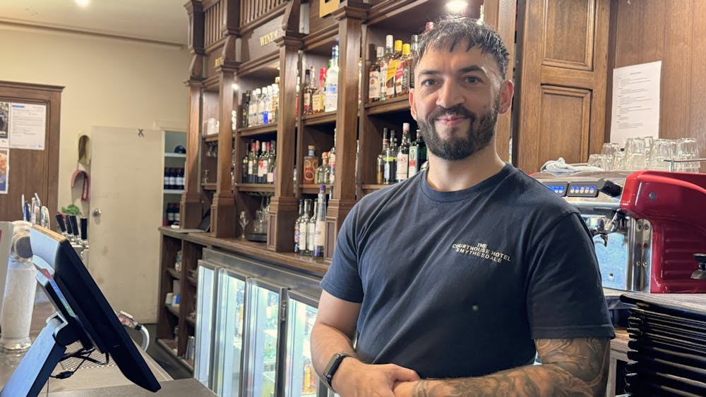 A staff member standing behind the bar at the Courthouse Hotel with shelves of spirits in the backgr