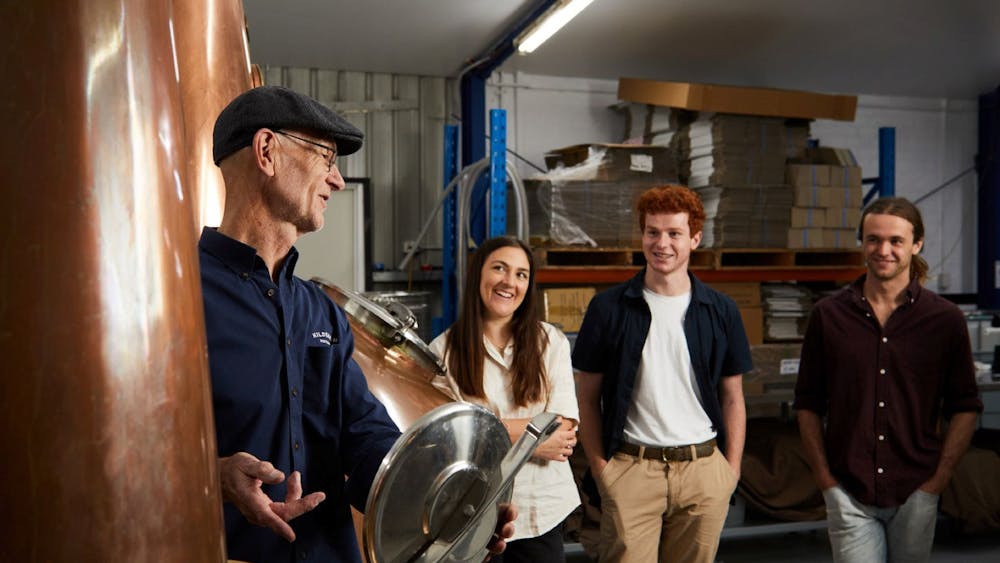 A small group of people watching a man as he holds the lid of a still open to explain its function