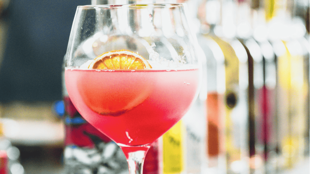 A pink cocktail in a large glass with a small dried orange piece floating in the glass
