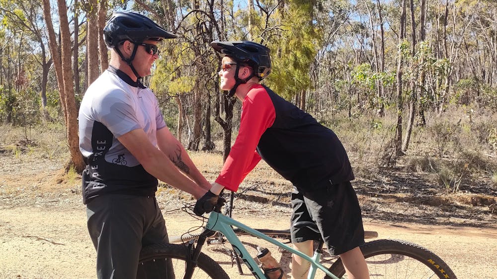 A man is holding onto the handle bars of a boy standing stationary on a mountain bike in the bush