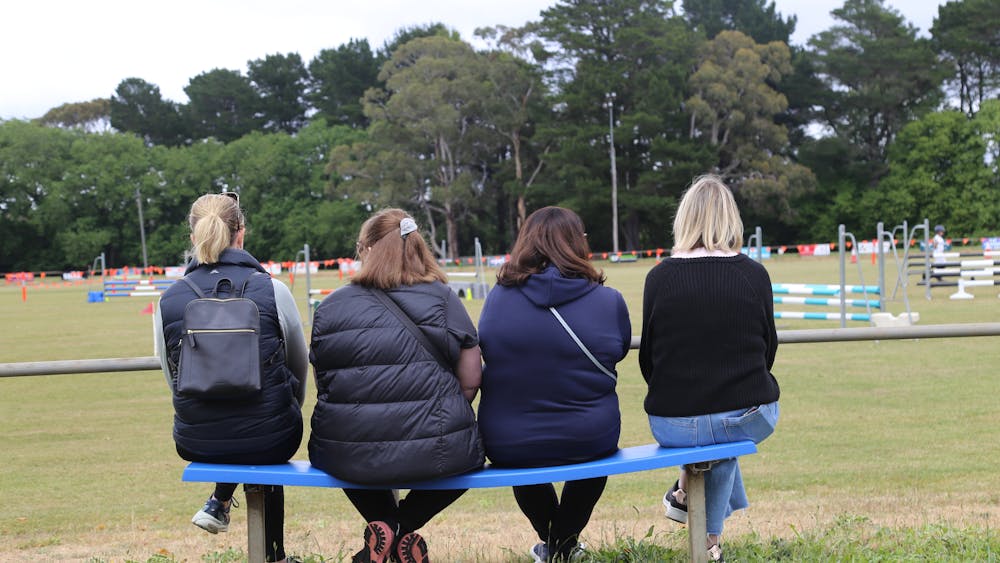 Backs of 4 women sitting on blue bench watching Show Jumping event