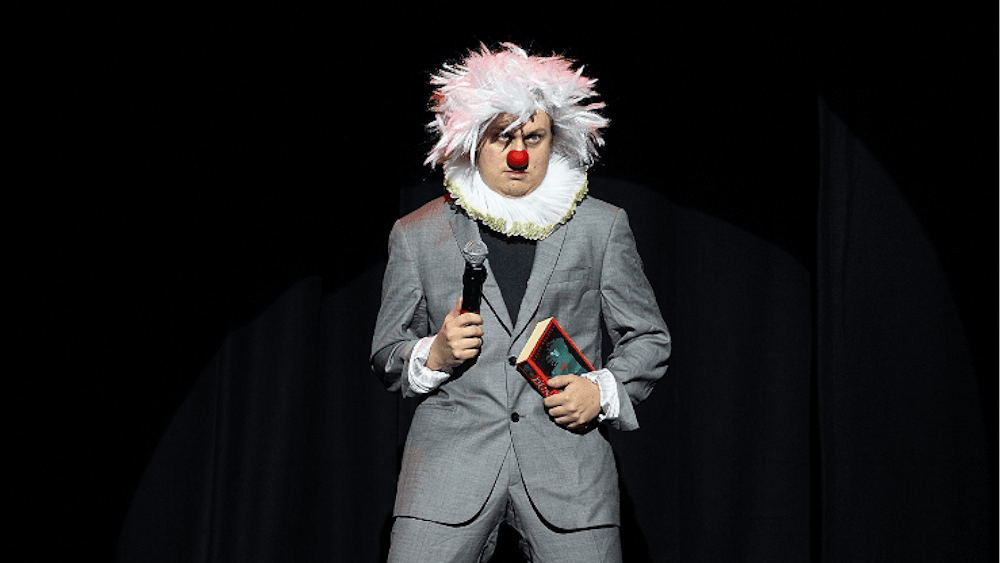 Artist in grey suit with white afro wig , red nose and white frilly color