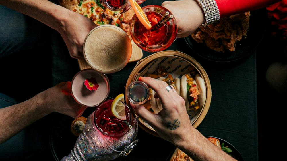 Overhead view of guests raising drinks around a table of bao, pizza and snacks.