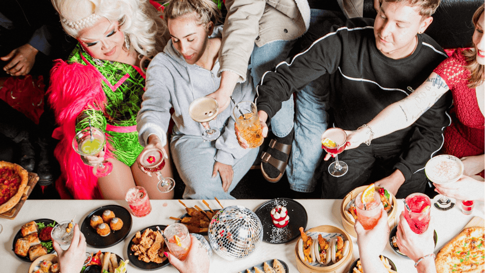 Guests raise cocktails around a table of shared food with a drag performer.